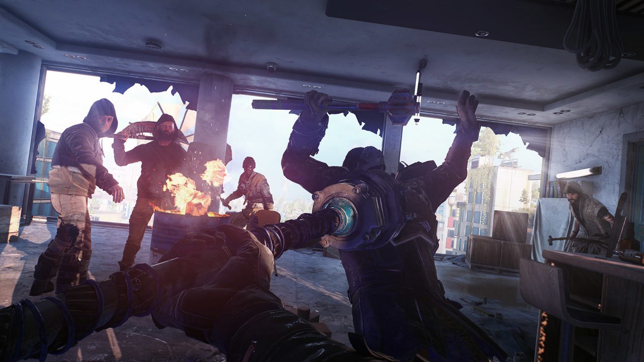 The main character battles vicious zombies in Dying Light 2 Stay Human. Men stand around an oil drum fire, and the wall is blown out. There is a desk to the right, and wooden crates in the background.