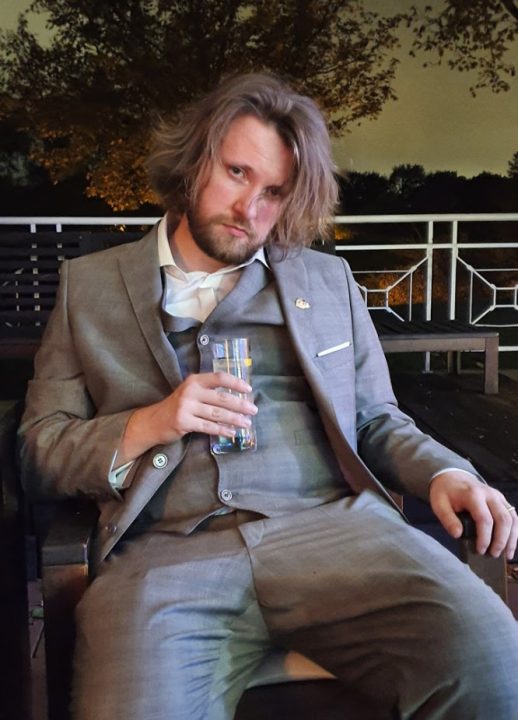 Matthias Linda slouching in a suit in a patio chair holding a tall glass of water, and looking tired