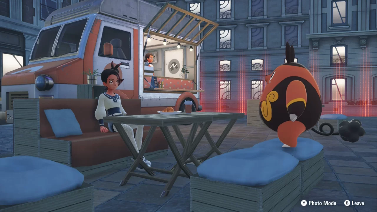 A Pokémon Legends: Z-A screenshot of the protagonist and a Pignite sitting outside around a table with a coffee and croissant. Behind them is a food truck.